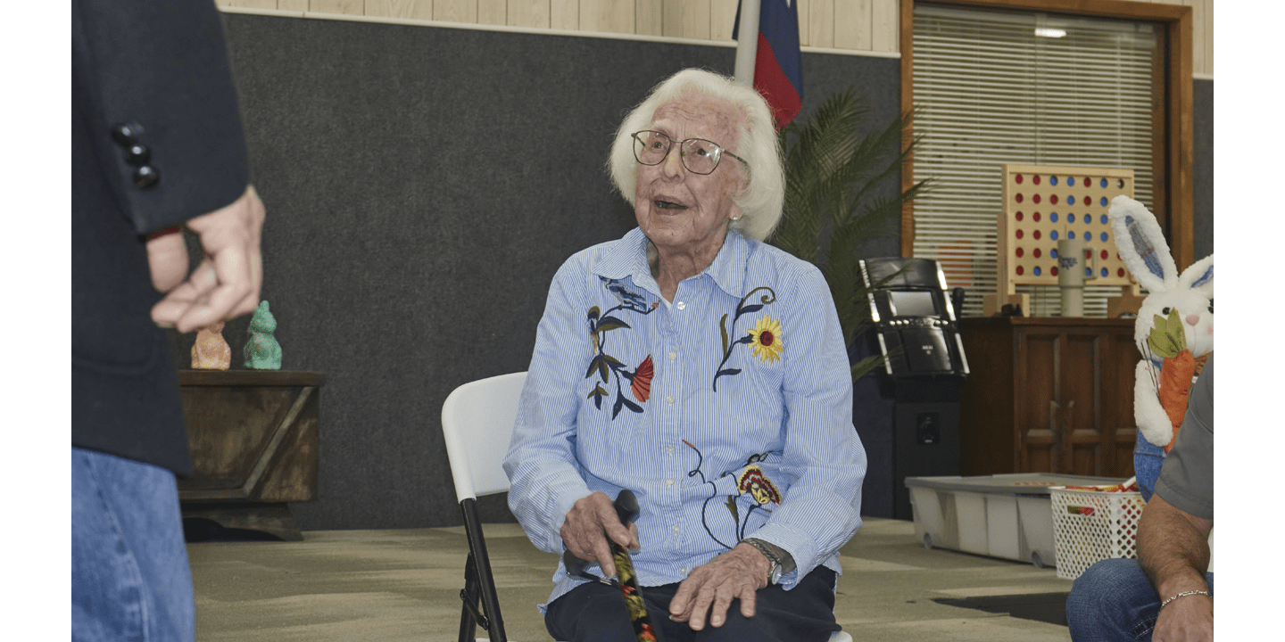 A lady with gray hair, glasses, and a blue and white striped shirt with flowers poses and talks to someone off camera, looking away from the camera.